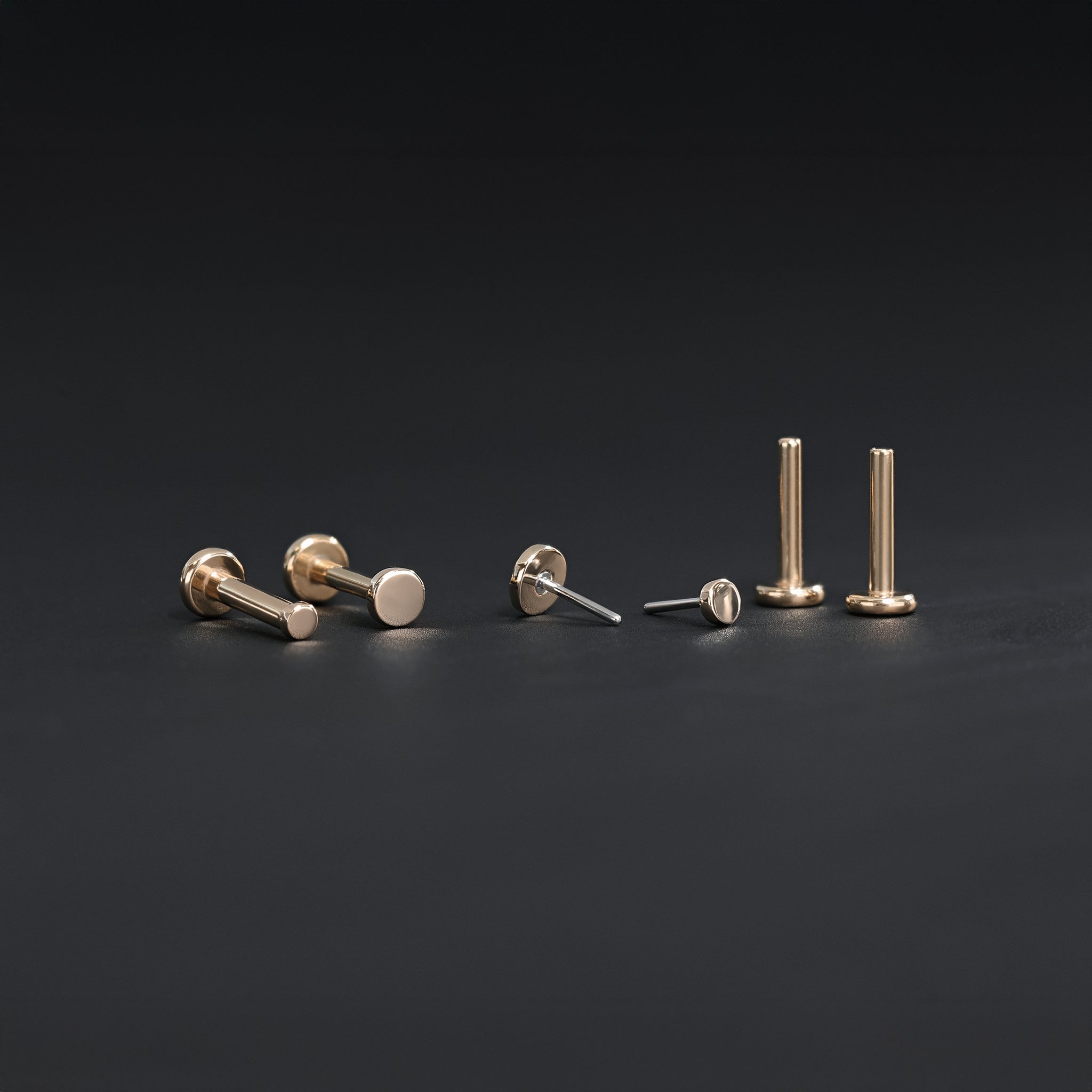 All four sizes 1.5mm, 2mm, 2.5mm and 3mm of 14K gold flat disc studs in rose gold displayed in different angles on black surface.