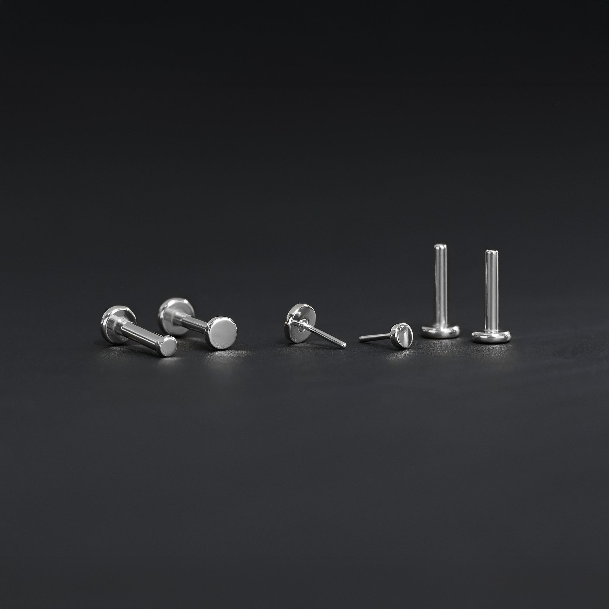 All four sizes 1.5mm, 2mm, 2.5mm and 3mm of 14K gold flat disc studs in white gold displayed in different angles on black surface.