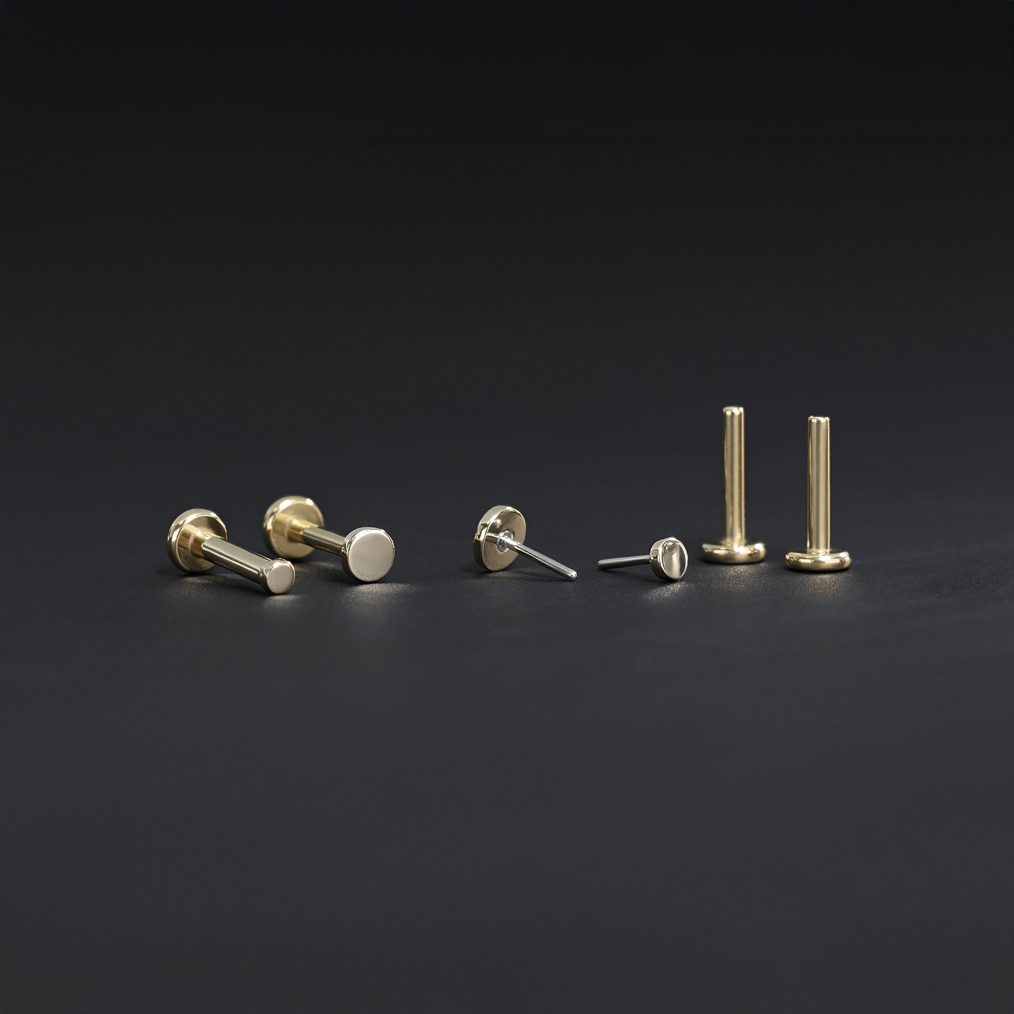 All four sizes 1.5mm, 2mm, 2.5mm and 3mm of 14K gold flat disc studs in yellow gold displayed in different angles on black surface.