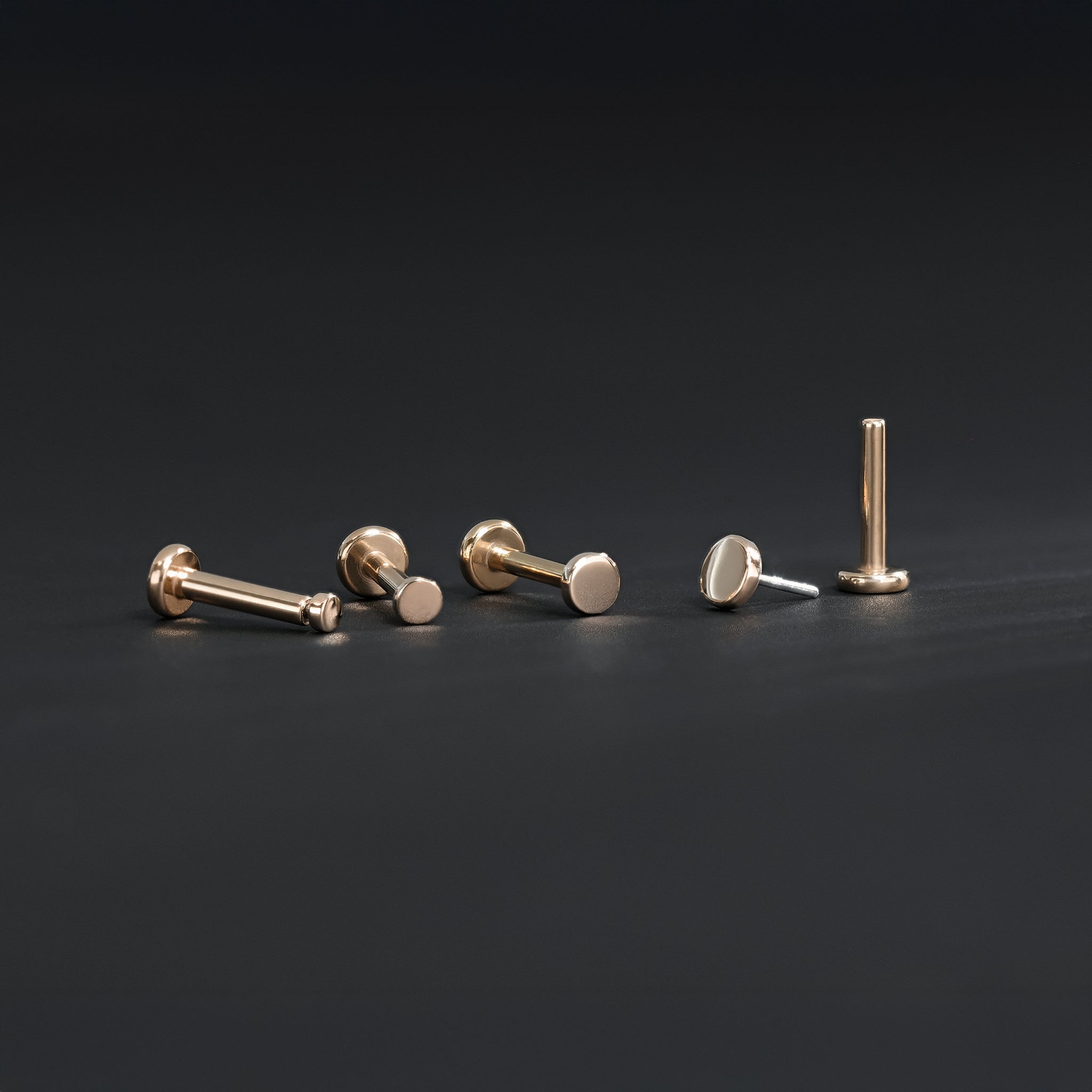 All four sizes 1.5mm, 2mm, 2.5mm and 3mm of 14K gold flat disc studs in rose gold displayed in different angles on black surface.