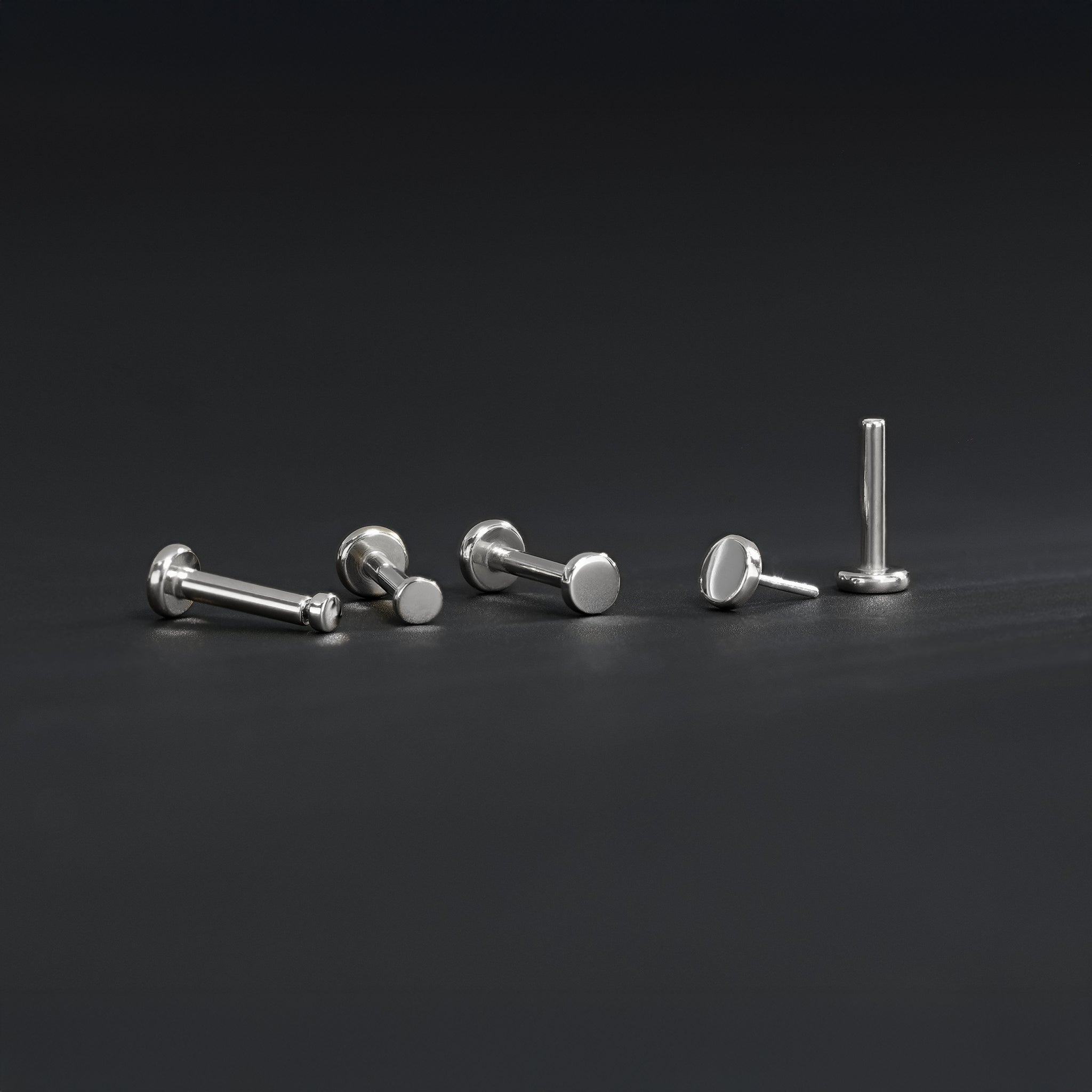 All four sizes 1.5mm, 2mm, 2.5mm and 3mm of 14K gold flat disc studs in white gold displayed in different angles on black surface.