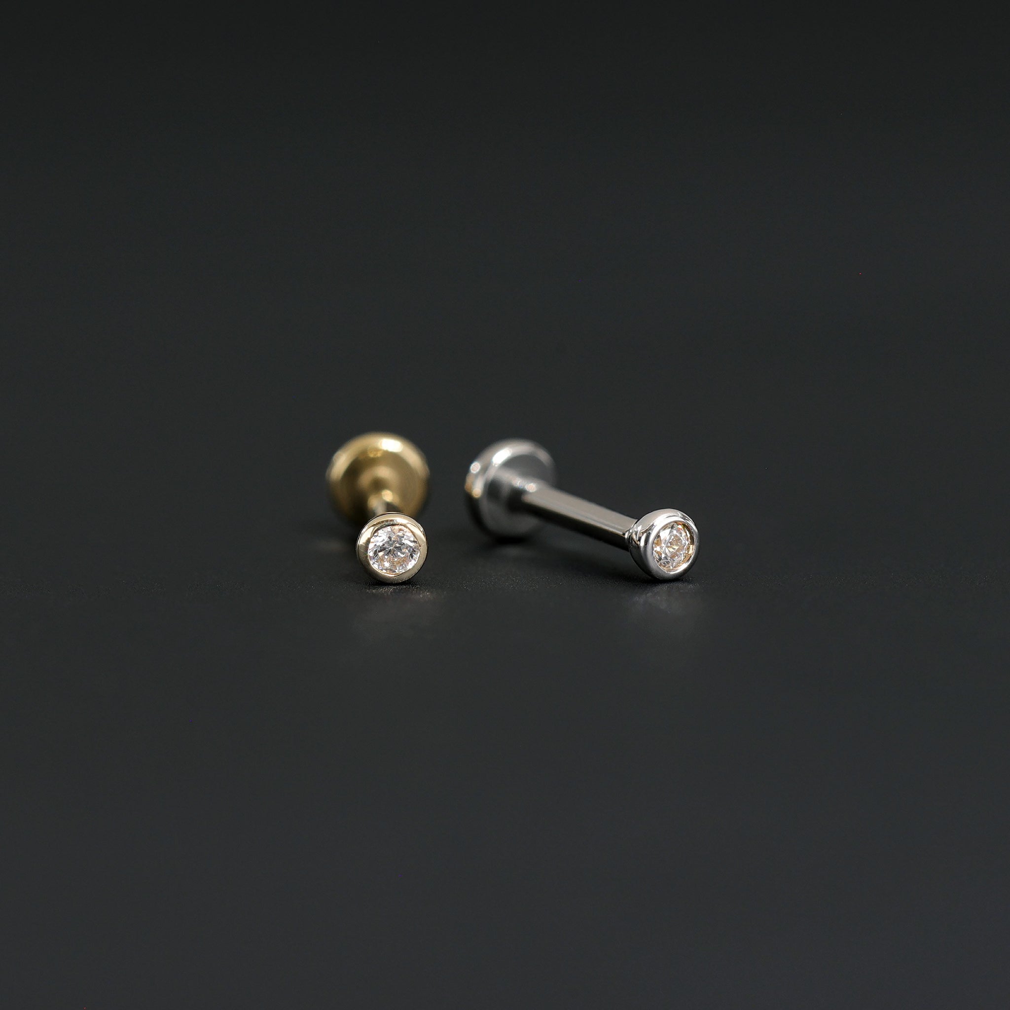 14K Gold Bezel Cubic Zirconia studs in yellow gold and white gold displayed on black surface, featuring its sparkles of Cubic Zirconia.