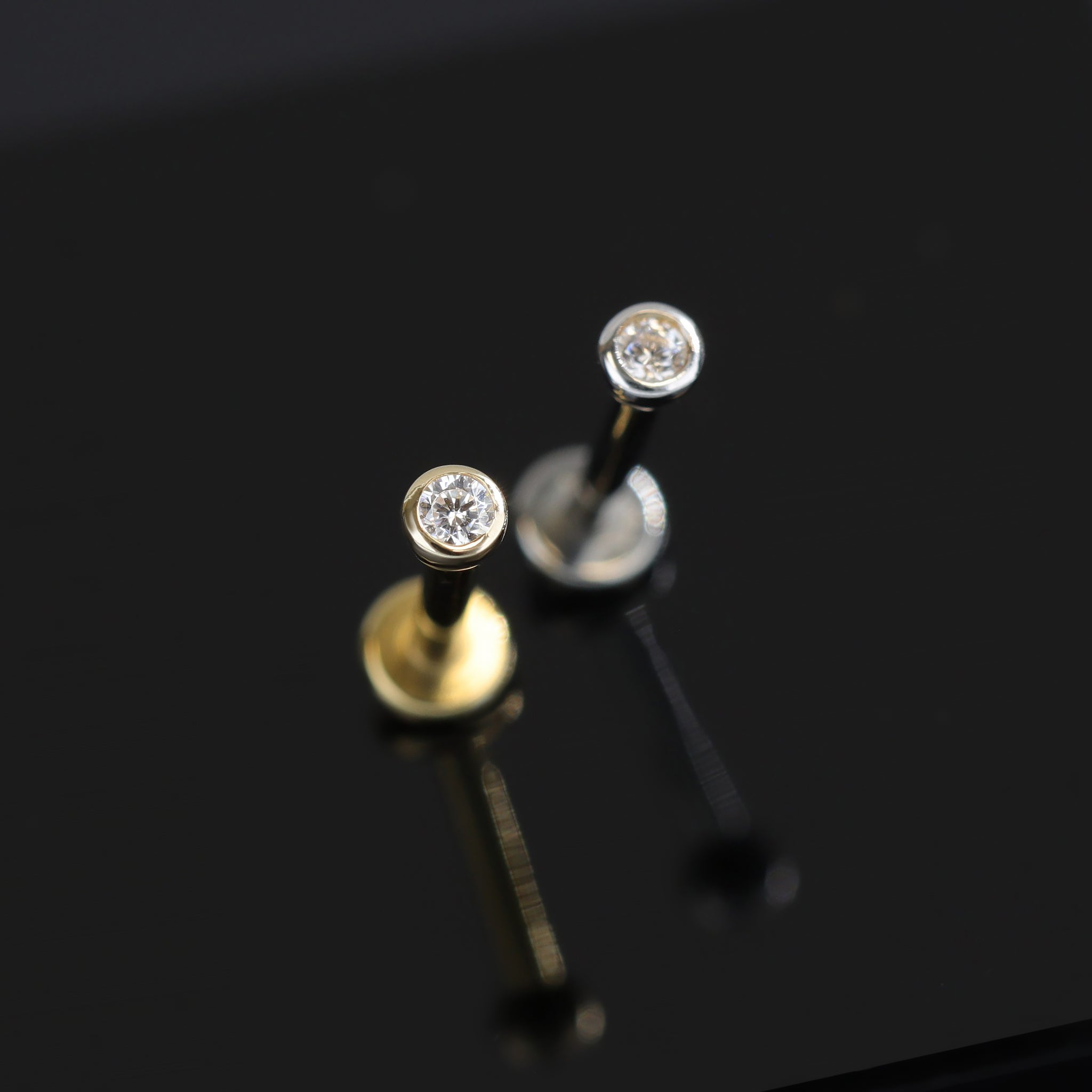 14K Gold Bezel Cubic Zirconia studs in yellow gold and white gold displayed on black surface, featuring its sparkles of Cubic Zirconia.