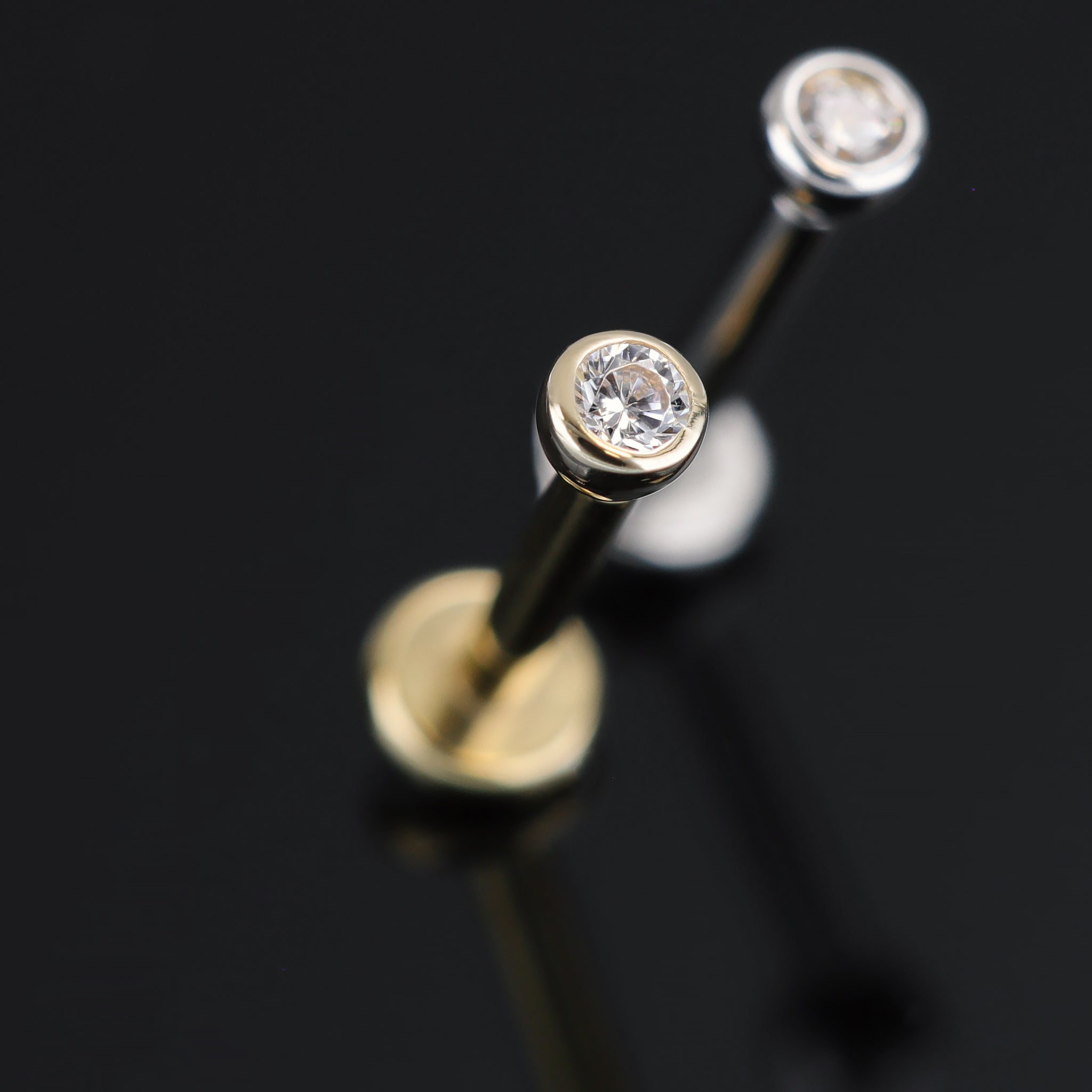 A close-up shot of 14K Gold Bezel Cubic Zirconia studs in yellow gold and white gold displayed on black surface, featuring its sparkles of Cubic Zirconia.