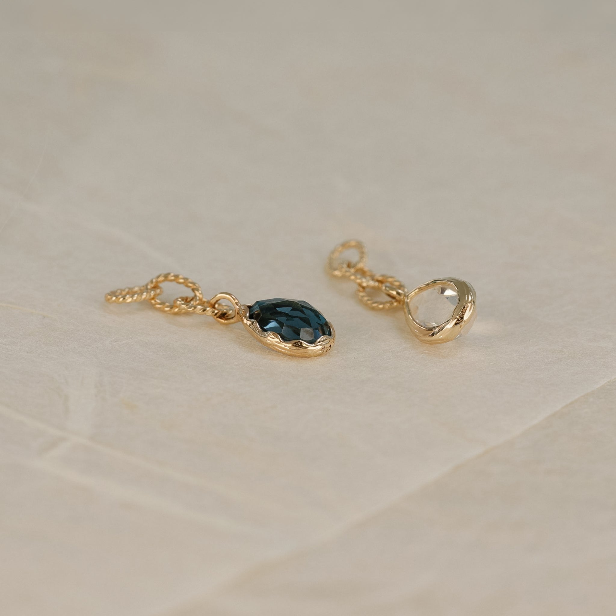 A teardrop charm of London Blue Topaz with gold twisted rope chain laid on white paper with Rainbow Moonstone Charm laid on the side upside down