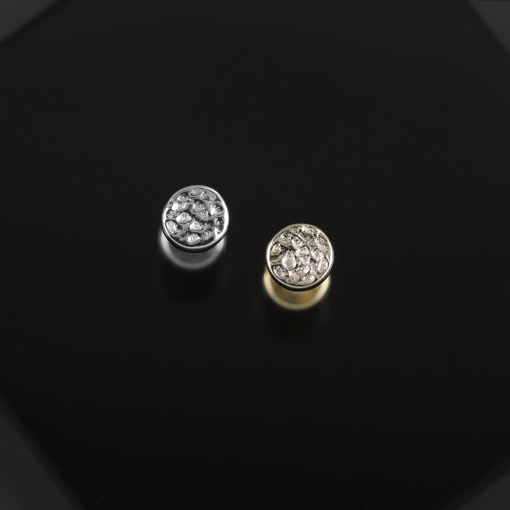 A close up shot of 3mm 14K Gold Hammered disc studs in yellow gold and white gold displayed on black surface, featuring its textured surface on a disc.